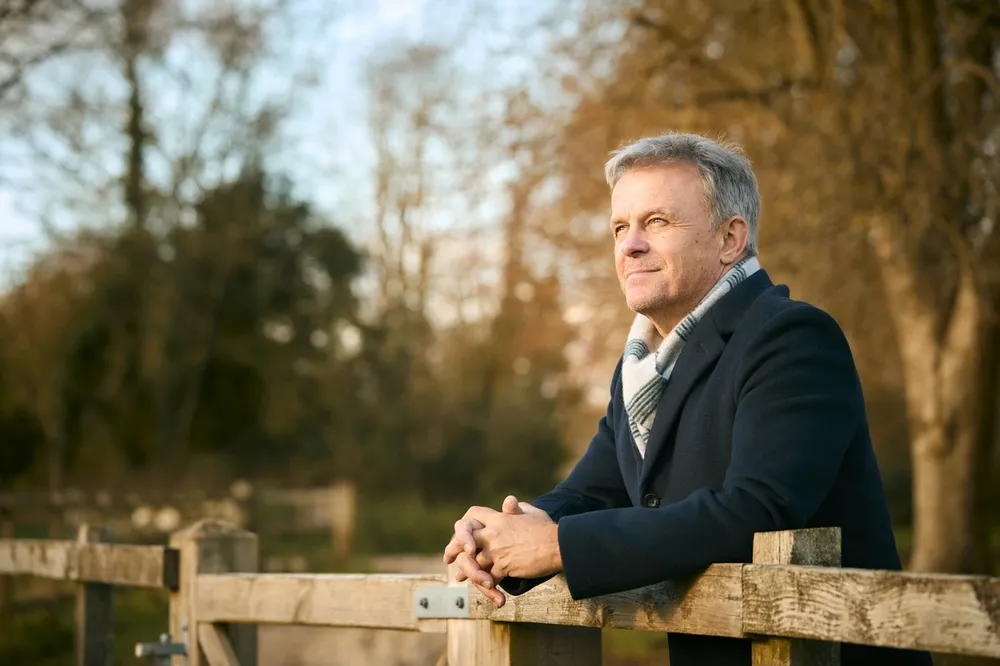 Smiling Senior Man Leaning On Wooden Fence On Walk Through Autumn Park Or Countryside