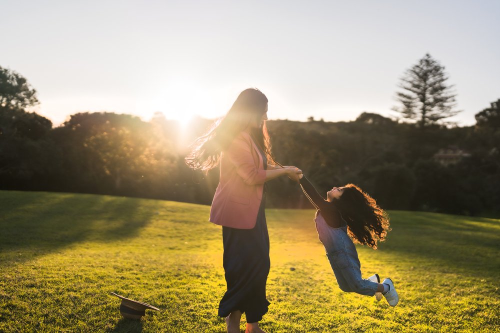 Woman swings daughter around by her arms in a grassy field at sunset