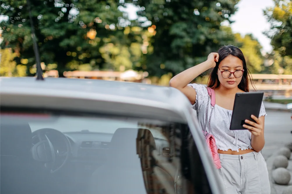 Young student holding an ipad leaning against a silver car