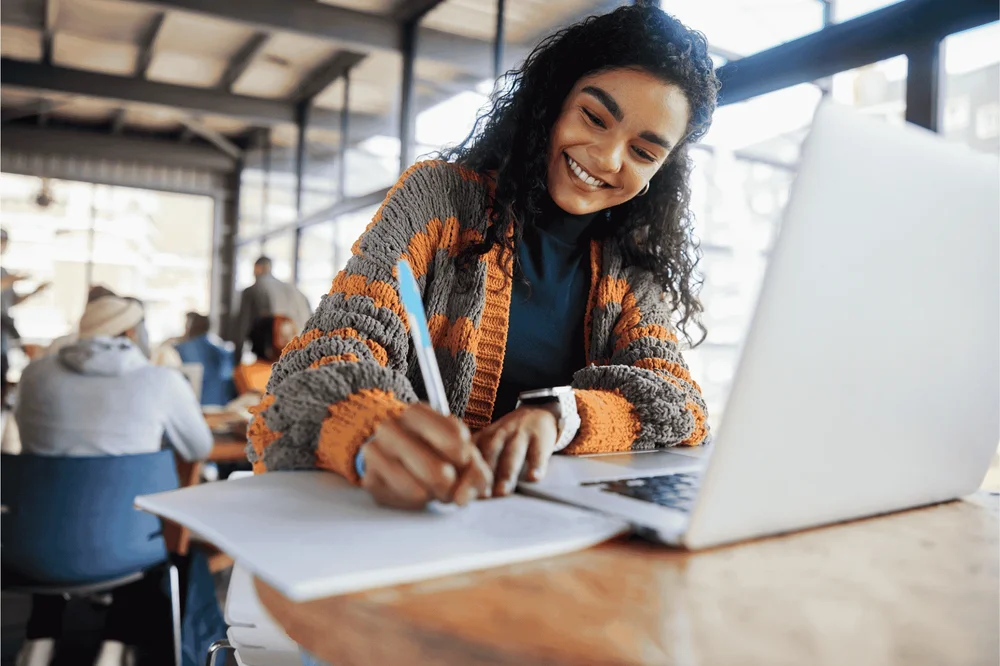 a young student smiling and writing notes with an open laptop